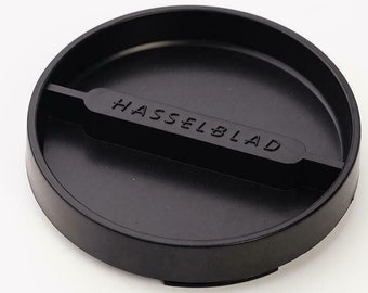 May include: A black camera lens cap with the word "HASSELBLAD" printed across the center. The cap is circular with a recessed interior and a small handle-like bar. The cap is made of a matte black material.