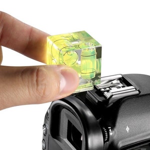May include: A clear, cube-shaped spirit level with yellow liquid inside is mounted on a black camera. The level is being held by a person's hand. The camera has a flash mount and various dials.