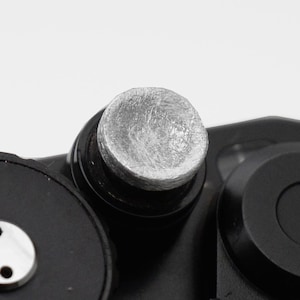 Könnte beinhalten: Nahaufnahme eines schwarzen Kameragehäuses mit einem silbernen Knopf und einem Zifferblatt mit weißen Zahlen und einer roten 60. Das Zifferblatt dient zur Einstellung der Verschlusszeit. Die Kamera hat einen schwarzen Auslöser.