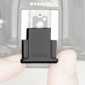 May include: A black metal flash cover for a camera. The cover is attached to the camera's hot shoe.