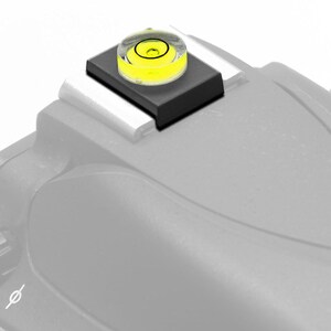 May include: A black camera hot shoe level with a clear bubble level. The bubble is yellow and has a black ring around it.