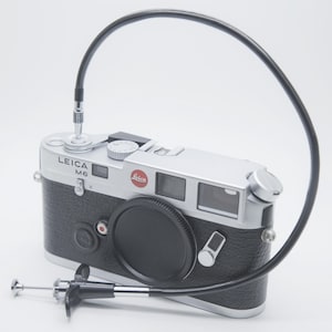 May include: A silver and black Leica M6 camera with a lens cap and a black cable release. The camera has a red Leica logo. The cable release is silver and black. The camera is on a white background.