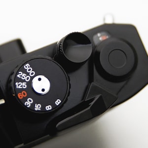Op de afbeelding: Close-up van een zwarte camera, met de nadruk op de wijzerplaten en bedieningselementen. De sluitertijdschijf is prominent aanwezig, met witte cijfers en een rode 60-markering. Andere zwarte wijzerplaten en knoppen zijn ook zichtbaar.
