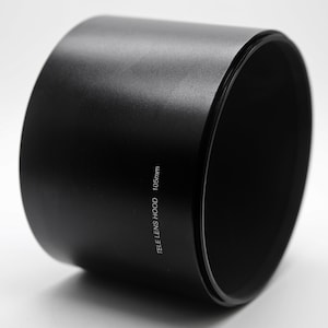 Può includere: Un paraluce per obiettivo nero, di forma cilindrica, con la scritta "TELE LENS HOOD 105mm" stampata sul lato. Il paraluce è progettato per ridurre i riflessi e migliorare la qualità dell'immagine. L'interno è nero opaco.
