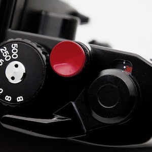 Può includere: Primo piano di una fotocamera nera con un pulsante rosso e un quadrante con numeri bianchi. Il design della fotocamera è elegante e minimalista, incentrato sulla funzionalità e sulla precisione. L'immagine evidenzia i componenti meccanici.