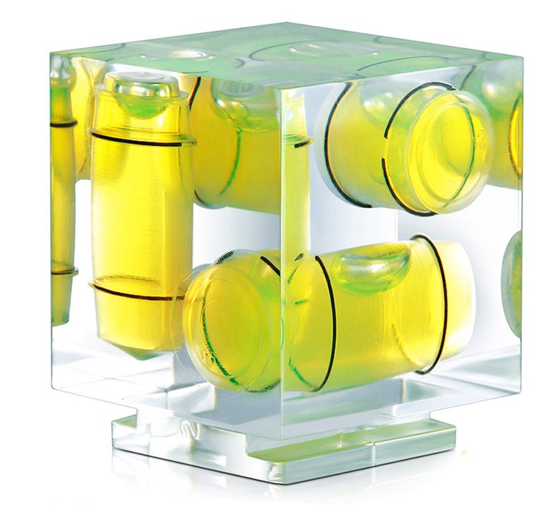 May include: A transparent, cube-shaped spirit level featuring multiple yellow and black bubble vials. The vials contain a bright yellow liquid. The level is designed for measuring horizontal and vertical alignment. Metric measurements.