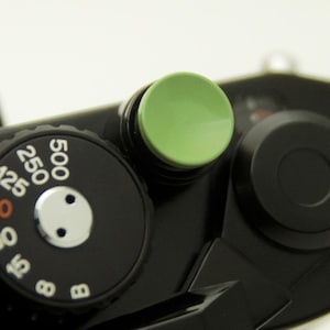 Könnte beinhalten: Nahaufnahme eines schwarzen Kameragehäuses mit einem grünen Knopf und einem Zifferblatt mit weißen Zahlen. Das Zifferblatt zeigt Verschlusszeiten von 8 bis 500 an. Das Kameradesign ist minimalistisch und konzentriert sich auf wesentliche Bedienelemente.