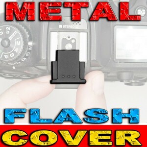 May include: A black metal flash cover for a camera. The cover is rectangular in shape with a small lip on the bottom. The cover is designed to protect the flash from damage.