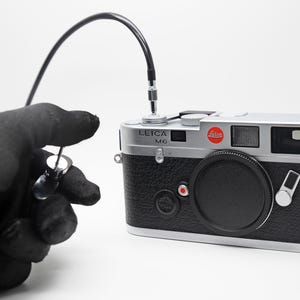 May include: A silver and black Leica M6 camera with a black lens cap and a red Leica logo. A black cable is connected to the camera. A black gloved hand holds a small, clear object.