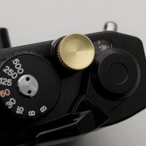 May include: Close-up of a black camera body with a gold-colored dial and black dials with white numbers. The camera has a sleek, minimalist design, with a focus on functionality and precision. The dials indicate settings for photography.
