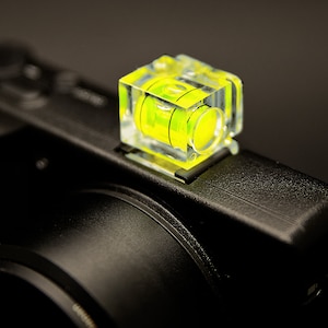 Può includere: Una livella a bolla cubica trasparente con una bolla giallo brillante, montata su una fotocamera nera. La livella è realizzata in plastica trasparente e si trova sulla slitta a contatto della fotocamera. Lo sfondo è scuro.