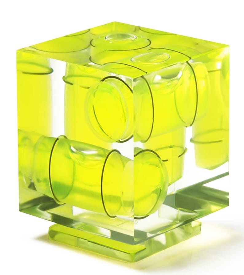 May include: A transparent, cube-shaped spirit level featuring multiple yellow bubble vials. The vials are positioned to show levelness across various axes. The level rests on a clear base, and the vials contain a bright yellow liquid.