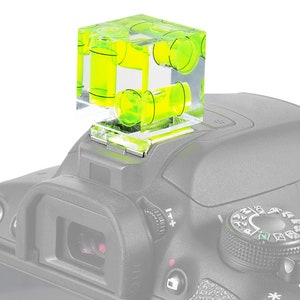 May include: A clear, cube-shaped spirit level with bright yellow liquid inside, mounted on top of a grey camera. The level has multiple bubble vials for precise alignment. The camera's controls are visible.