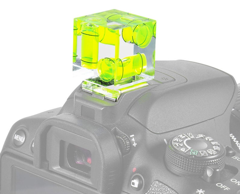 May include: A clear, cube-shaped spirit level with bright yellow liquid inside, mounted on top of a grey camera. The level has multiple bubble vials for precise alignment. The camera's controls are visible.