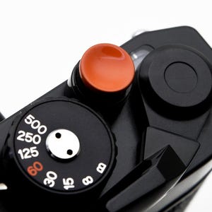 May include: Close-up of a black vintage camera with an orange shutter button and a black dial with white numbers. The dial indicates shutter speeds from 8 to 500. The camera's design is classic and minimalist.