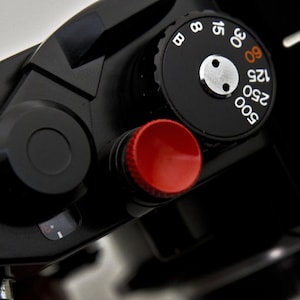 Può includere: Primo piano di un corpo macchina nero con un pulsante rosso e un quadrante nero. Il quadrante presenta numeri bianchi e un centro argentato. Il design della fotocamera è elegante e moderno, con un focus sulla funzionalità e precisione.