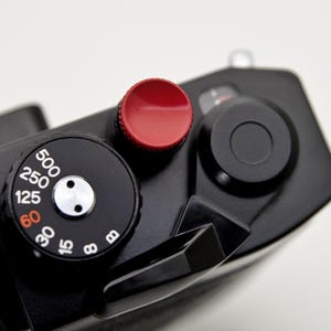 May include: Close-up of a black camera body with a red shutter button and a black dial with white numbers. The dial indicates various shutter speeds, including 500, 250, 125, and 60. The camera has a sleek, minimalist design.