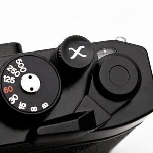 Può includere: Primo piano della parte superiore di una fotocamera nera, che evidenzia i quadranti e i controlli. Il quadrante della velocità mostra numeri bianchi, che vanno da 8 a 500. È visibile anche un pulsante nero con un simbolo "X" bianco.