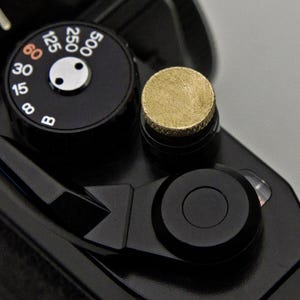 Può includere: Primo piano di un corpo macchina nero con un pulsante color oro e un quadrante nero con numeri bianchi. Il quadrante indica i tempi di posa da 8 a 500. Sono visibili anche una leva nera e un pulsante nero.
