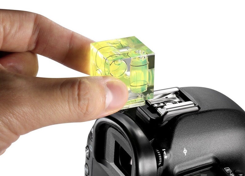 May include: A clear, cube-shaped spirit level with yellow liquid inside is mounted on a black camera. The level is being held by a person's hand. The camera has a flash mount and various dials.