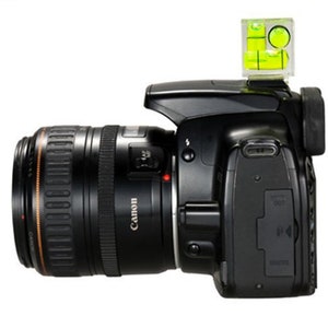 May include: Black Canon DSLR camera with a lens attached. A clear spirit level with yellow liquid is mounted on top. The lens has the word "Canon" printed on it.
