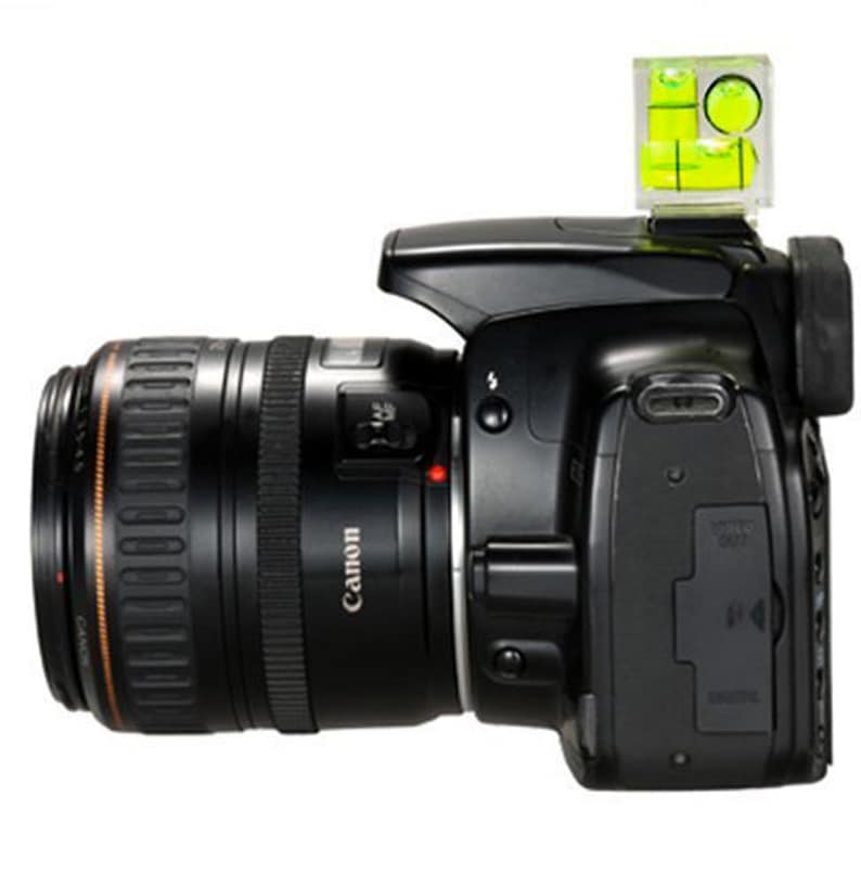 May include: Black Canon DSLR camera with a lens attached. A clear spirit level with yellow liquid is mounted on top. The lens has the word "Canon" printed on it.