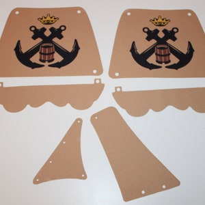 Könnte beinhalten: Ein Set aus sieben beigefarbenen Filzteilen für ein Piratenschiff-Spielset. Die Teile umfassen zwei große rechteckige Teile mit einem schwarzen Anker- und Fassdesign, zwei kleinere rechteckige Teile und drei gebogene Teile.