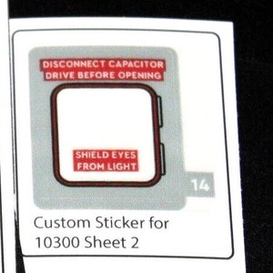 Può includere: Un adesivo personalizzato bianco con un design rettangolare e un bordo rosso scuro. L'adesivo riporta il testo "DISCONNECT CAPACITOR DRIVE BEFORE OPENING" e "SHIELD EYES FROM LIGHT". È presente anche il testo "Custom Sticker for 10300 Sheet 2".