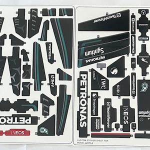 以下が含まれることがあります： 黒、白、緑のロゴとテキストが書かれた、モデルカー用のステッカーシート。ロゴには、ペトロナス、イネオス、チームビューワー、IWC、スナップドラゴン、シンチウムが含まれています。