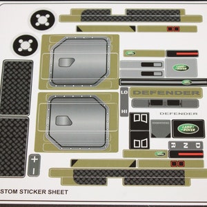 Könnte beinhalten: Ein Bogen mit individuellen Aufklebern mit Land Rover Defender-Designs. Die Aufkleber umfassen Türverkleidungen, Kühlergrills und andere Fahrzeugdetails in Grau-, Schwarz- und Olivgrüntönen. Der Text "DEFENDER" ist sichtbar.