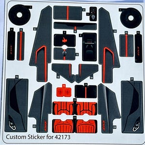 May include: A sheet of black and red stickers for a custom car model. The stickers include various shapes and designs, including a car logo, seats, and side panels. The text "Custom Sticker for 42173" is printed at the bottom of the sheet.