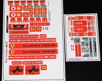 Pode incluir: Uma folha de adesivos para um caminhão modelo, com os logotipos da Scuderia Ferrari, Iveco, Fiat, Bridgestone, Vodafone, AMD, Olympus, Brembo, Mahle, SKF, Acer, Momo e Beta. A folha é dividida em duas seções, etiquetadas 1/2 e 2/2. A folha é branca com acentos vermelhos e pretos.