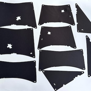Könnte beinhalten: Eine Ansammlung schwarzer Papierauschnitte in verschiedenen Formen, darunter Rechtecke und Dreiecke, einige mit sternförmigen Löchern und ausgefransten Kanten. Die Teile haben kleine Löcher, wahrscheinlich zur Montage, auf weißem Hintergrund.