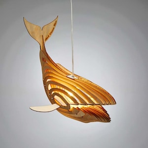 Op de afbeelding: Een houten hanglamp in de vorm van een walvis met een gloeilamp erin. De walvis is gemaakt van dun, gelaagd hout en heeft een natuurlijke houtkleur. De staart van de walvis is omhoog gebogen en de bek van de walvis is open. De lamp hangt aan een wit snoer.