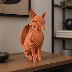 May include: A terracotta-colored fox sculpture, crafted from layered wooden slats, sits on a wooden surface. The fox is depicted in a seated position, with its ears perked up and tail curved. The sculpture is a decorative item.