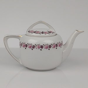 May include: White porcelain teapot with a floral design featuring pink and purple roses. The teapot has a lid with a handle and a spout.