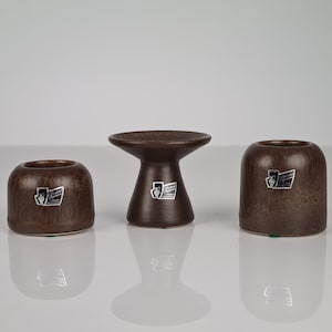 Puede incluir: Tres candelabros de cerámica marrón con un logotipo blanco. Los candelabros están en fila sobre una superficie blanca.