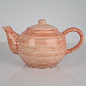 May include: A light pink ceramic teapot with a striped pattern. The teapot has a lid and a handle.