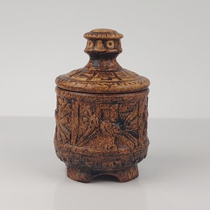 Puede incluir: Un pequeño tarro de cerámica marrón con tapa. El tarro tiene tallas intrincadas y una base redondeada.