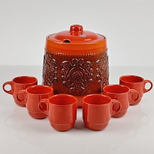 Puede incluir: Un juego de tazas de cerámica naranja y una jarra a juego con un diseño floral en relieve. La jarra tiene una tapa con un pequeño pico.