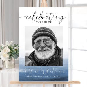 May include: A memorial sign on an easel with a black and white portrait of a man wearing glasses and a knit cap. The sign reads "Celebrating the Life of Arthur Whitmore" with dates April 1st 1944 - July 17th 2025.