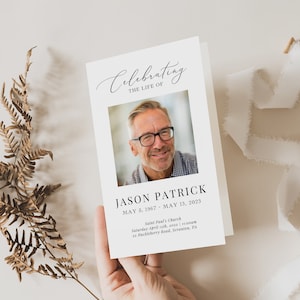 May include: A folded memorial card featuring a photograph of a man wearing glasses. The text reads "Celebrating the Life of" and "Jason Patrick, May 5, 1967 - May 13, 2023." A sprig of dried foliage and a white ribbon are also present.