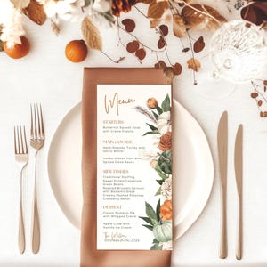 May include: A printed menu card titled Menu with sections for Starters, Main Course, Side Dishes, and Dessert, decorated with watercolor illustrations of pumpkins and autumn foliage, resting on a brown napkin and a white dinner plate.