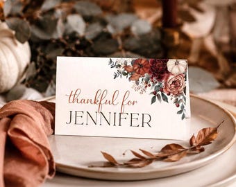 May include: A white rectangular place card featuring a floral arrangement of dark red and orange flowers with green leaves in the top right corner. The text reads "thankful for" in a script font above the name "JENNIFER" in a bold serif font.