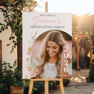 May include: A wooden easel displays a graduation party sign. The sign reads "Welcome to Sienna's Graduation Party" with a photo of a smiling woman. Floral accents and the year 2026 are also featured. The sign is from Sunny Hills High School.