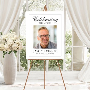 May include: A white memorial sign with black text that reads "Celebrating the Life of Jason Patrick 01.05.1967 - 13.03.2022". A photo of a man with glasses is in the center of the sign.