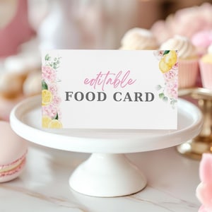 May include: A white food card with the text "editable FOOD CARD" in pink and gray. The card is decorated with watercolor illustrations of lemons, pink roses, and greenery. The card sits on a white, tiered cake stand.