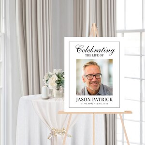 May include: White memorial sign with black text that reads "Celebrating the Life of Jason Patrick 01.05.1967 - 13.05.2022". A photo of a man with glasses is in the center of the sign.