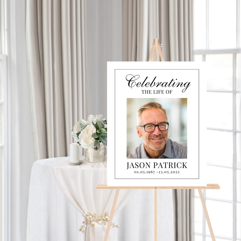 May include: White memorial sign with black text that reads "Celebrating the Life of Jason Patrick 01.05.1967 - 13.05.2022". A photo of a man with glasses is in the center of the sign.