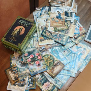 Op de afbeelding: Een verzameling tarotkaarten met gouden randen en aquarel illustraties, verspreid rond een groene doos. Op de doos staat "Whispers of the Tao" en "Tarot of Tao De Jing". De kaarten tonen afbeeldingen van de natuur, figuren en symbolen.